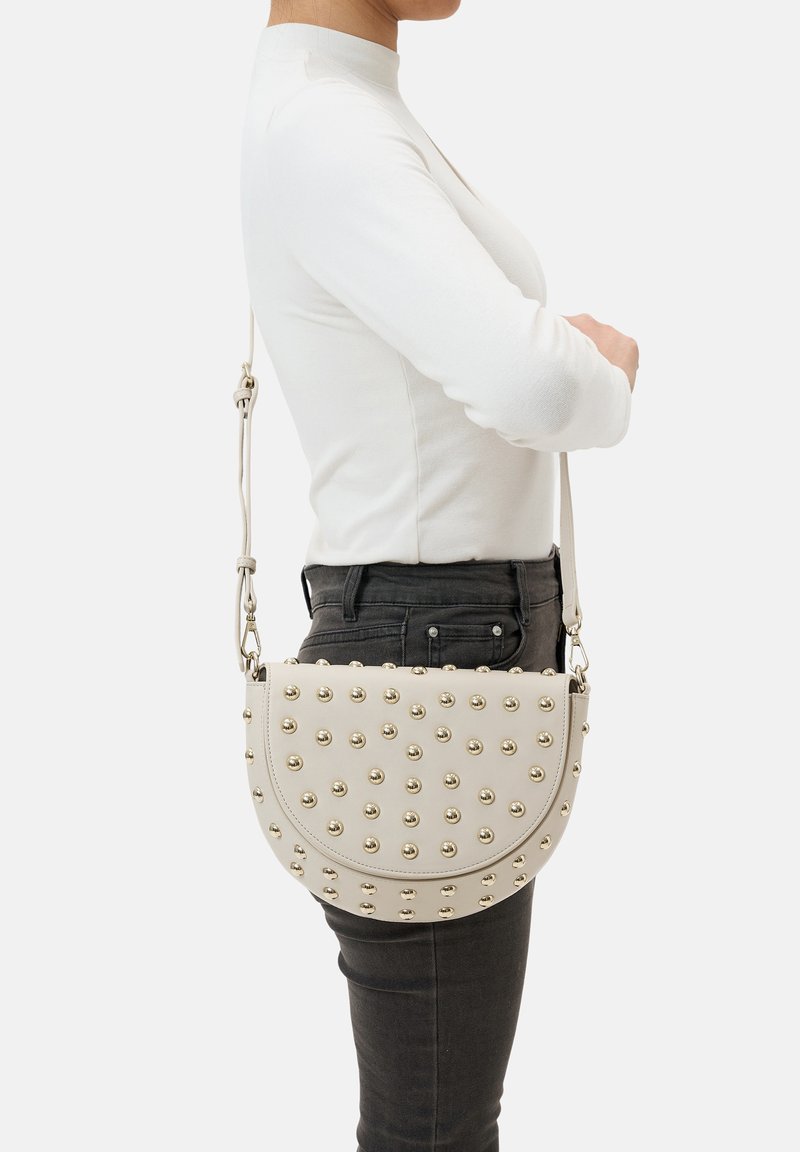Sac crossbody beige avec des clous dorés, en forme de demi-cercle, texture lisse, bandoulière ajustable, porté sur le côté contre un jean noir.