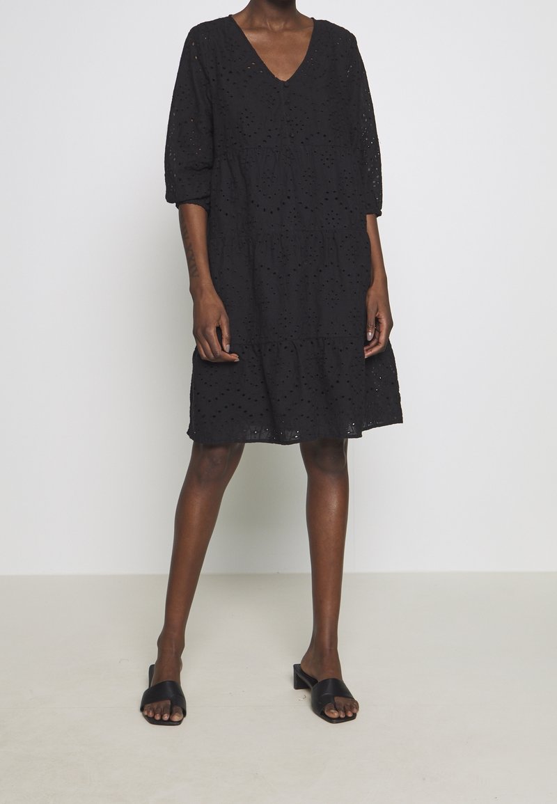 Femme portant une robe noire à œillets longueur genou avec des manches trois-quarts et des sandales noires ouvertes à l’avant, sur un fond uni.