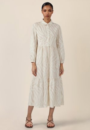 Frau trägt ein langärmeliges, knopfgeschlossenes Midikleid mit dezentem beigen Zebramuster und braunen Riemchensandalen, steht vor einem neutralen Hintergrund.