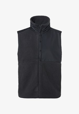 Schwarze Weste mit hohem Kragen, die eine Kombination aus glatten und strukturierten Materialien zeigt. Vorderknöpfe und Markenzeichen an der unteren Seite.