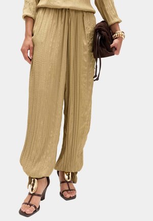 Kvinde iført beige teksturerede bukser med vide ben og snøre ved anklerne, brune sandaler med hæl og stropper, holder en mørkebrun clutch og et guldarmbånd.
