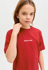 Fiatal, világosbarna hajú személy piros pólóban, amelyen az "Originals Studio" felirat olvasható, enyhén megérinti a gallérját, egyszerű fehér háttér előtt.