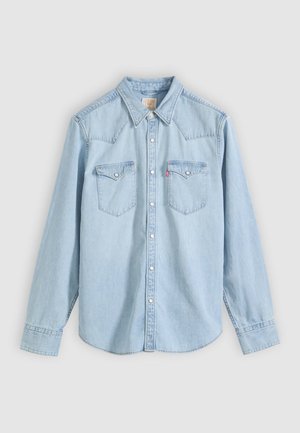 Ανοιχτό μπλε πουκάμισο από τζιν με μακριά μανίκια, κουμπιά μπροστά, δύο τσέπες στο στήθος με καπάκι και κουμπιά, καθώς και μυτερό γιακά σε μονόχρωμο φόντο.