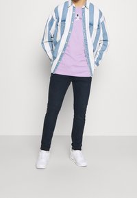 Person som bär vita sneakers, mörka slim fit jeans, en lila grafisk t-shirt och en uppknäppt blå-vitrandig denimskjorta.