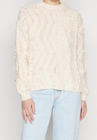 Vaaleanbeige neule, jossa on teksturoitu, aaltoileva kuvio. Varustettu ribbireunuksilla hihansuissa ja helmassa, sekä löysällä leikkauksella. Yhdistetty vaaleansinisiin farkkuihin.