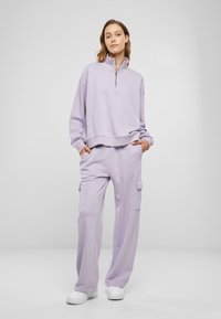 Helllila Fleece-Zip-Hoodie mit hohem Kragen, kombiniert mit lockeren, passenden Hosen mit Cargo-Taschen. Weiße Sneakers.