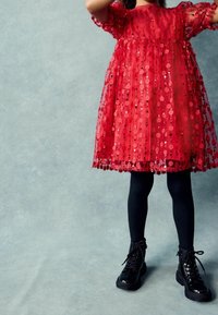 Robe en tulle rouge ornée de sequins floraux, avec manches courtes bouffantes et taille froncée. Portée avec des bottines noires et des leggings.