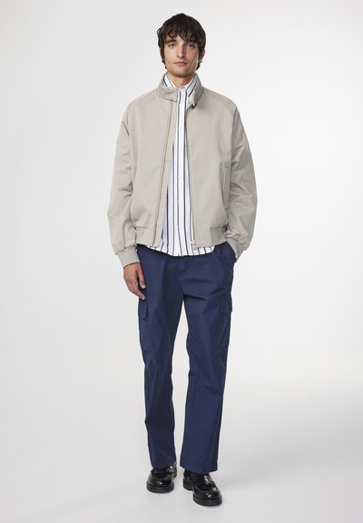 Giacca bomber beige con colletto rialzato, chiusura a zip, pantaloni cargo blu navy, camicia bianca e blu a righe, e scarpe nere. Texture lisce, design minimale.