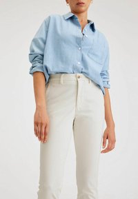 Camicia azzurro chiaro con taschino sul petto, maniche arrotolate, abbinata a pantaloni aderenti color crema con dettaglio a un bottone.