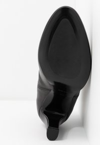 Scarpa nera in pelle con tacco alto che mostra la suola liscia con una piccola punta del tacco su uno sfondo bianco.