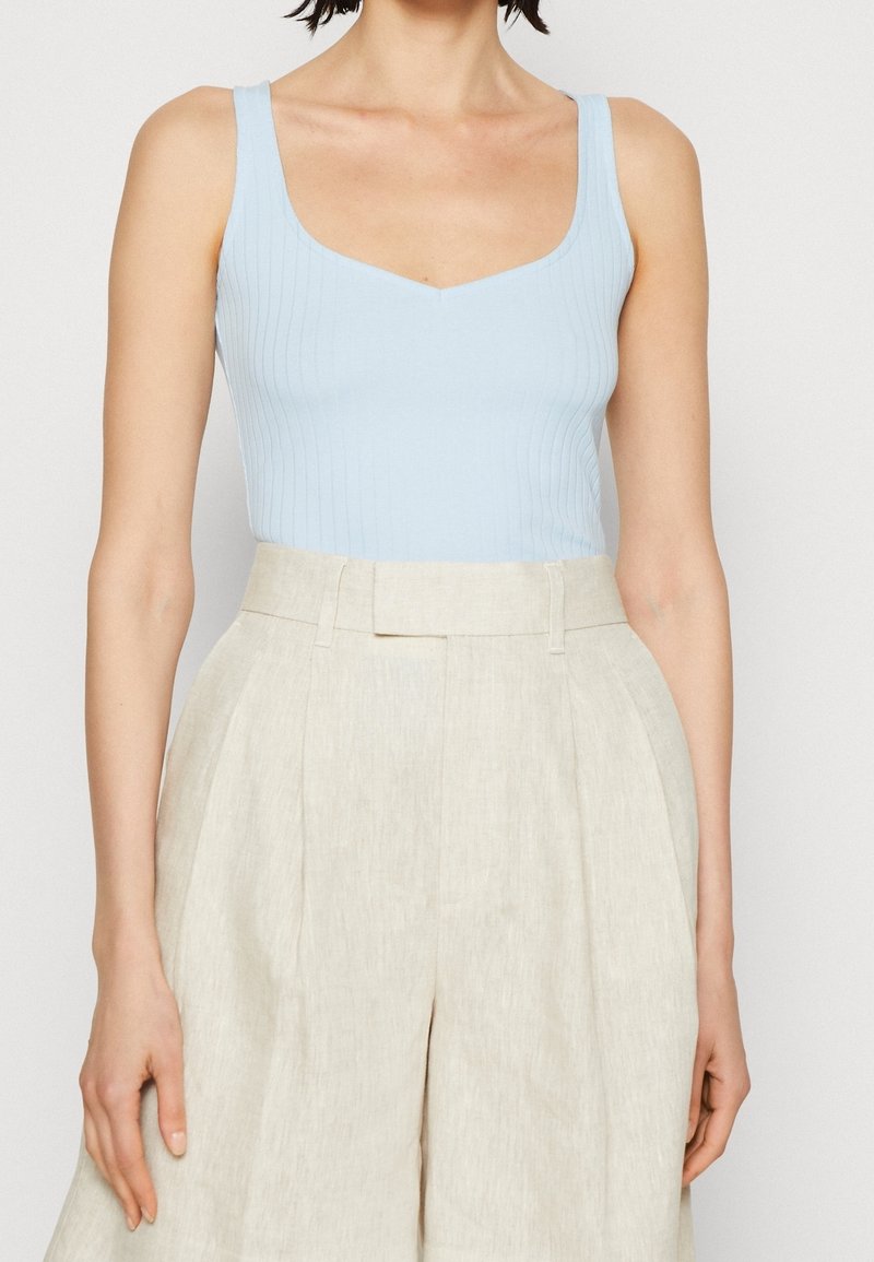 Débardeur bleu clair côtelé avec un col en V, associé à un pantalon beige taille haute avec des plis et une coupe ajustée. Texture lisse dans l'ensemble.