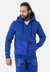 Blaue Zip-Hoodie mit Kängurutasche, kontrastierendem strukturiertem Vorderteil, Kapuze mit Zugband und markiertem Lederpatch-Detail.