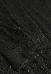 Obleka s črnimi bleščicami, prikazana pod podrobno povečavo teksture tkanine, manšetom z gumbom in naborki.