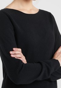 Pull en tricot noir avec une texture douce, encolure bateau et manches longues, affichant une coupe décontractée et un design minimaliste.