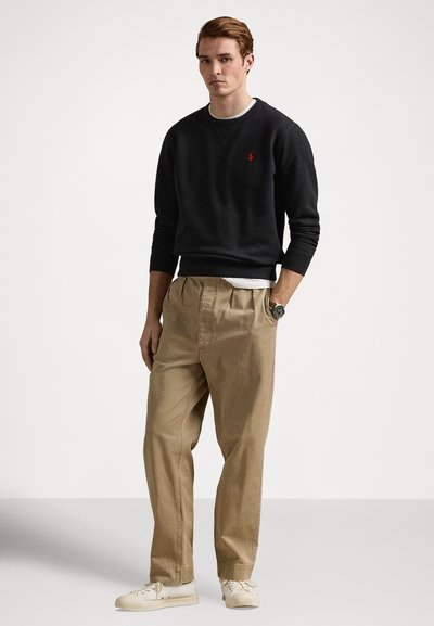 Μαύρο φούτερ με μικρό κόκκινο λογότυπο, συνδυασμένο με μπεζ παντελόνι χαλαρής εφαρμογής. Άσπρη μπλούζα από κάτω, φορεμένη με άσπρα αθλητικά παπούτσια.