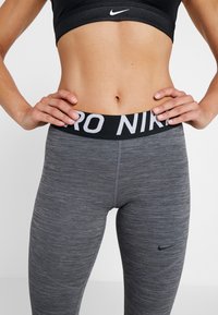 Leggings Nike Pro en gris foncé, dotés d'une large taille noire avec le texte "PRO NIK" en blanc. Tissu lisse et extensible avec une texture chinée.