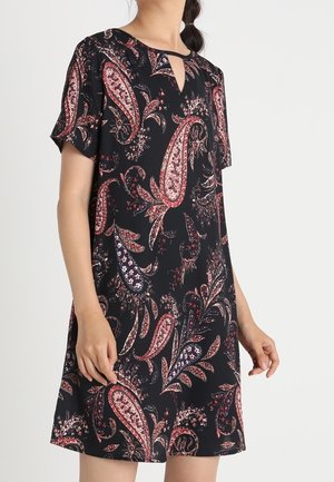 Mujer vistiendo un vestido corto negro con patrones de cachemira en rojo y blanco, y mangas cortas, sobre un fondo liso.