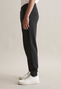 Schwarze Jogginghose mit elastischem Bund, seitlichen Taschen und schmal zulaufenden Beinen. Kombiniert mit weißen Turnschuhen mit strukturierten Sohlen.