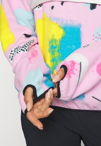 Mains avec de longs ongles sortant des trous pour le pouce d’un sweat polaire coloré à motifs roses, jaunes, bleus et noirs.