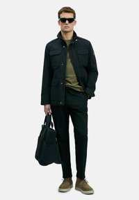 Man in een zwarte jas, olijfgroen geribbeld sweater, zwarte broek, tan suède schoenen, zwarte zonnebril, houdend een zwarte gewatteerde totebag.