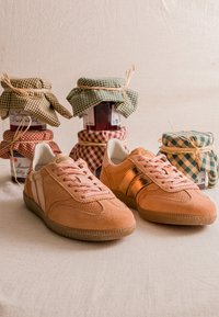 Paire de baskets en daim marron clair avec des lacets roses devant des bocaux en verre empilés, recouverts d’un tissu à carreaux et attachés avec de la ficelle.