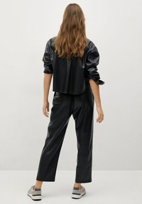 Chaqueta corta de cuero sintético negro con mangas abullonadas, combinada con pantalones de tiro alto a juego y zapatillas grises. Modelo de espaldas.