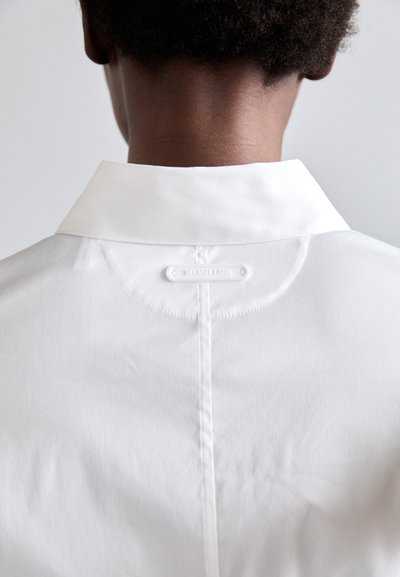 Fehér ing klasszikus gallérral, sima textúrával és hátsó márkajelzéssel. Egyszerű design, tiszta vonalakkal és minimális részletezéssel.