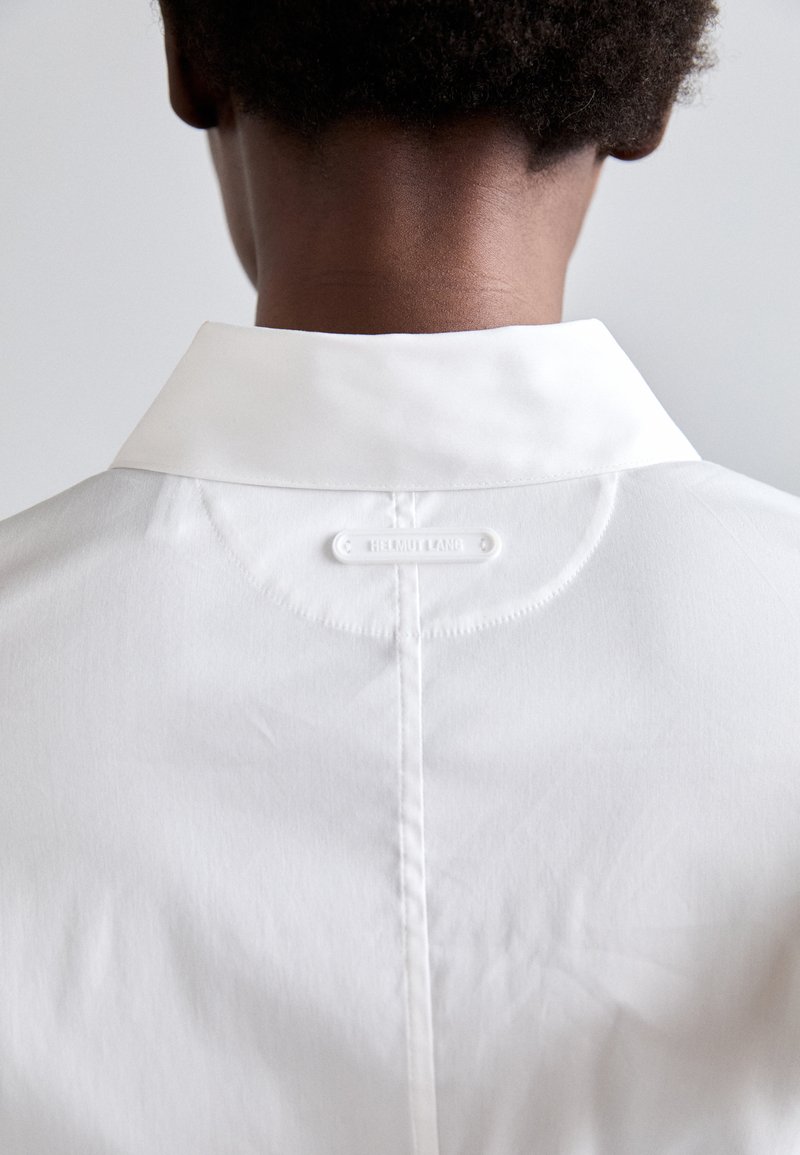 Hvid skjorte med klassisk krave, glat tekstur og et mærke-tag på bagsiden. Enkel design med rene linjer og minimal detaljer.