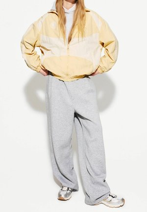 Person iført en gul og hvid lynlåstrøje, løse grå joggingbukser og sølv- og hvid sneakers, stående med hænderne på hofterne.