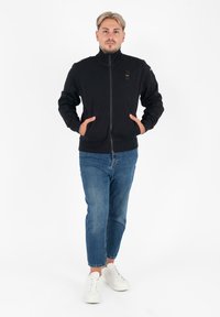Zwarte zip-up jas met een hoge kraag, twee voorzakken en logoaccenten. Gecombineerd met blauwe denim en witte sneakers. Eenvoudig ontwerp.