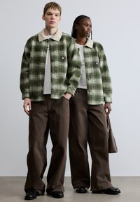 Groen geblokt shacket met een beige sherpa-kraag, gecombineerd met bruine broek met wijde pijpen. Beiden dragen witte T-shirts, met een bruine tasdetail.