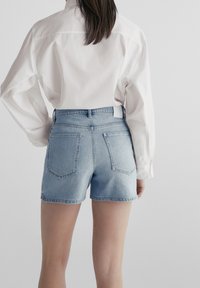 Jasnoniebieskie szorty z denimu z wysokim stanem, z pięcioma kieszeniami i postrzępionym dołem. Modelka ma na sobie białą koszulę z długim rękawem.