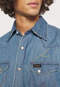 Camicetta in denim di colore azzurro chiaro, colletto a punta, ricami dorati, due taschini sul petto con bottoni in perlina e logo Wrangler.