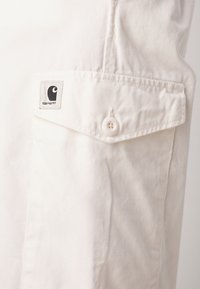 Chemise en coton blanc avec une poche sur la poitrine dotée d'un bouton et d'une étiquette logo noire. Texture lisse et design simple avec des accents minimaux.