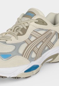Μπεζ και γκρι παπούτσι τρεξίματος Asics με ανοιχτό καφέ ρίγες, μπλε λεπτομέρειες και λευκή σόλα, εμφανίζοντας λεπτομέρειες από δίχτυ και κορδόνια.