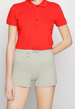 Mujer con una camiseta polo de manga corta roja brillante y pantalones cortos de punto gris claro con cordón, de pie frente a un fondo simple.