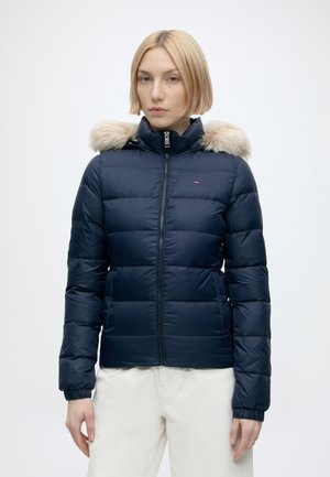 Jonge vrouw met kort blond haar die een donkerblauwe donsjas met bontkraag en een witte broek draagt, staand tegen een egale achtergrond.
