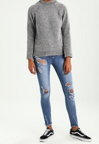 Grauer Strickpullover mit gerippter Textur, kombiniert mit blauen, verwaschenen Skinny-Jeans mit mehreren Rissen und schwarzen Canvas-Sneakers.