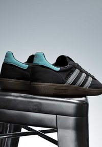 Baskets Adidas noires avec des rayures grises et des languettes turquoises au talon affichant les logos Mercedes AMG, placées sur un tabouret métallique sur fond gris.