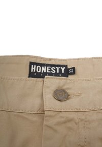 Beige bomuldsbukser med en metalknap og syningdetaljer. Mærket siger "HONESTY" med størrelsesmærke "32." Glat tekstur, afslappet design.