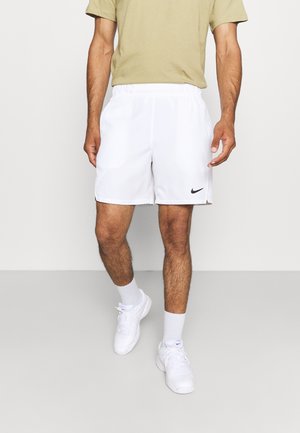 Mann trägt weiße Nike-Sportshorts, weiße Socken und weiße Turnschuhe, steht drinnen vor einem einfachen hellen Hintergrund.