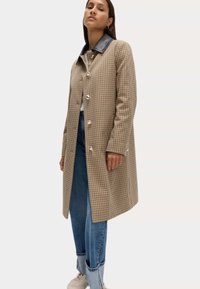 Cappotto a scacchi marrone con colletto in pelle nera, dotato di chiusura a bottone e maniche lunghe, indossato sopra jeans blu e sneakers chiare.