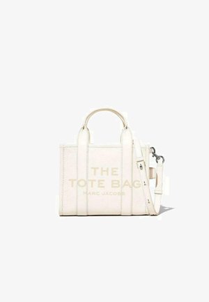 Witte leren schoudertas met twee handvatten, een afneembare schouderband en een gebosstere tekst die zegt "DE TOTE BAG MARC JACOBS." Deze tas heeft een gladde textuur.