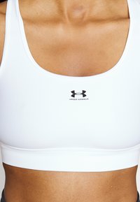 Witte sportbeha gemaakt van gladde, rekbare stof. Heeft een ronde halslijn en een smalle onderband, met een zwart logo in het midden van de voorkant.
