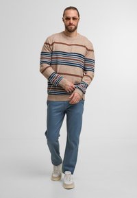 Beżowy sweter dzianinowy w poziome brązowe i niebieskie paski, zestawiony z niebieskimi spodniami i beżowymi trampkami, charakteryzujący się luźnym krojem i ściągaczami.