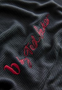 Černá vroubkovaná tkanina s texturovanými pruhy, která obsahuje výrazný červený výšivku "by Ted Baker." Detailní pohled zdůrazňuje materiál a zpracování.