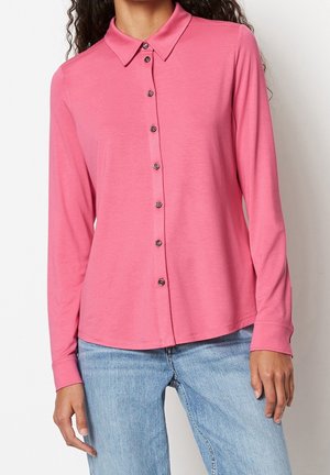 Kobieta w różowej koszuli na guziki z długim rękawem i kołnierzykiem oraz jasnoniebieskich dżinsach, stojąca na białym tle.