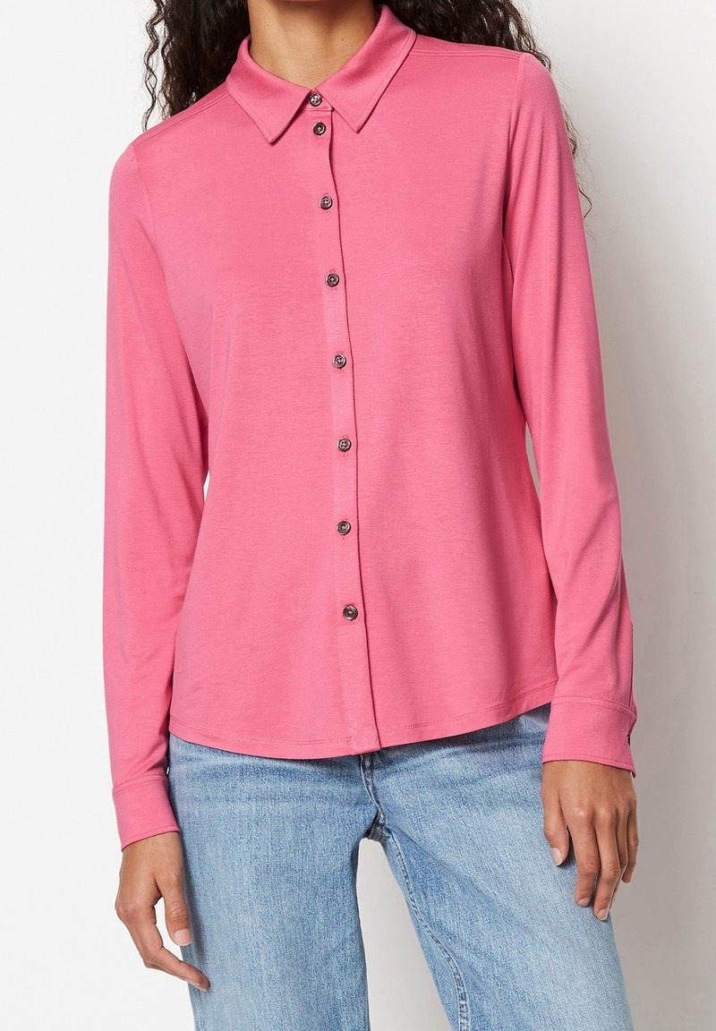 Femme portant une chemise rose à manches longues avec col et boutons ainsi qu’un jean en denim bleu clair, debout devant un fond blanc.
