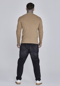 Bež dugih rukava, pripijena; tamne traperice širokog kroja, do gležnjeva; bijele tenisice s tamnoplavim đonom; vidljive tetovaže na vratu.