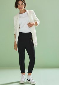 Sacou alb cu dungi peste o cămașă albă, pantaloni negri tapered cu buzunar cu fermoar și adidași albi cu un logo negru. Fundal verde deschis.